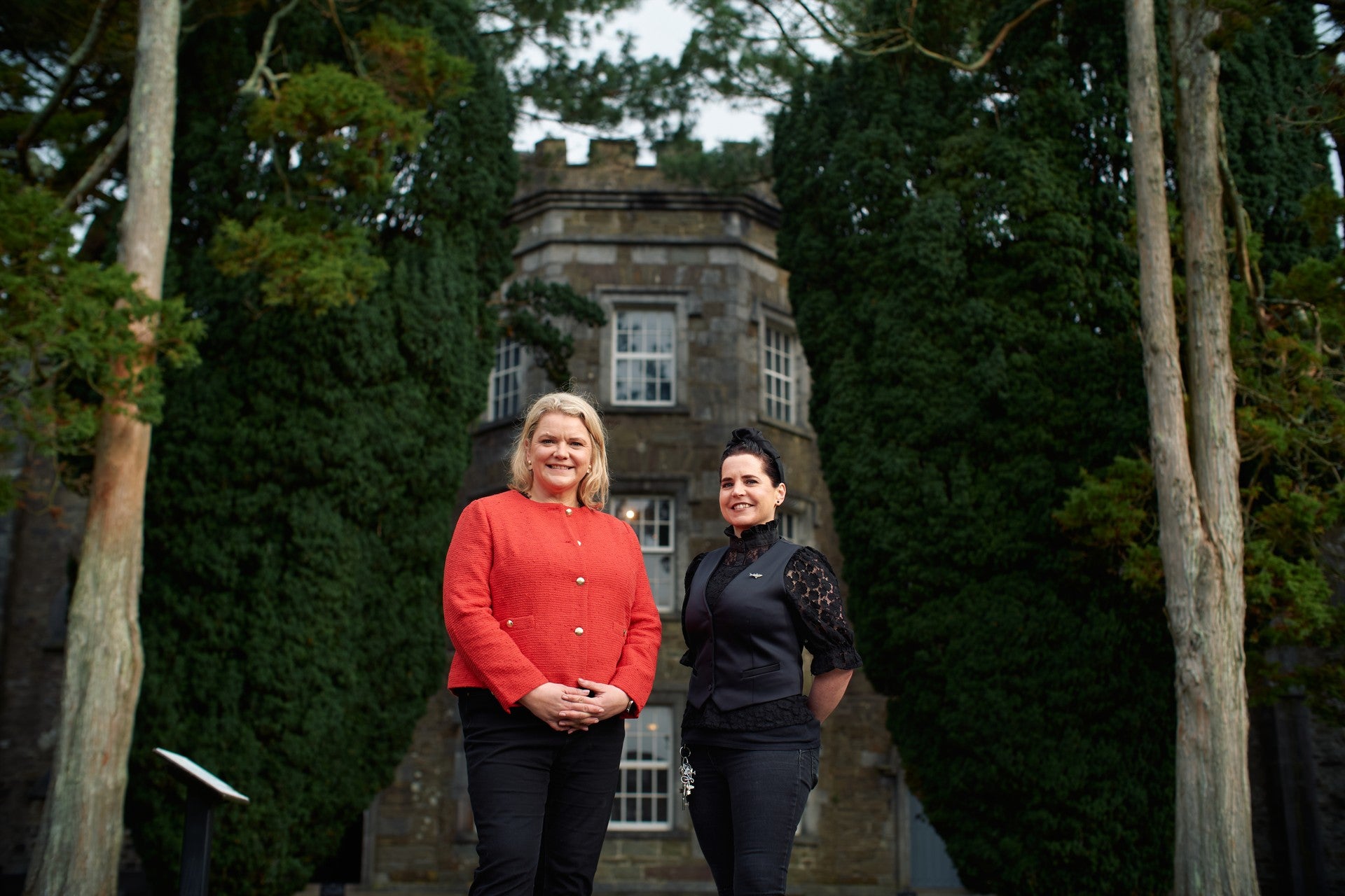 Two people standing outdoors in front of a historic stone manor house surrounded by tall trees