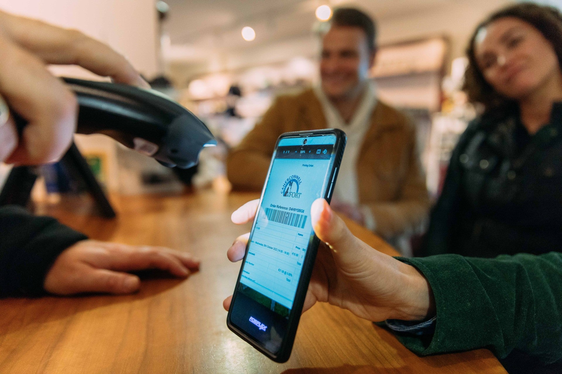 Person scanning a digital booking confirmation on a smartphone at a venue counter, with customers in the background