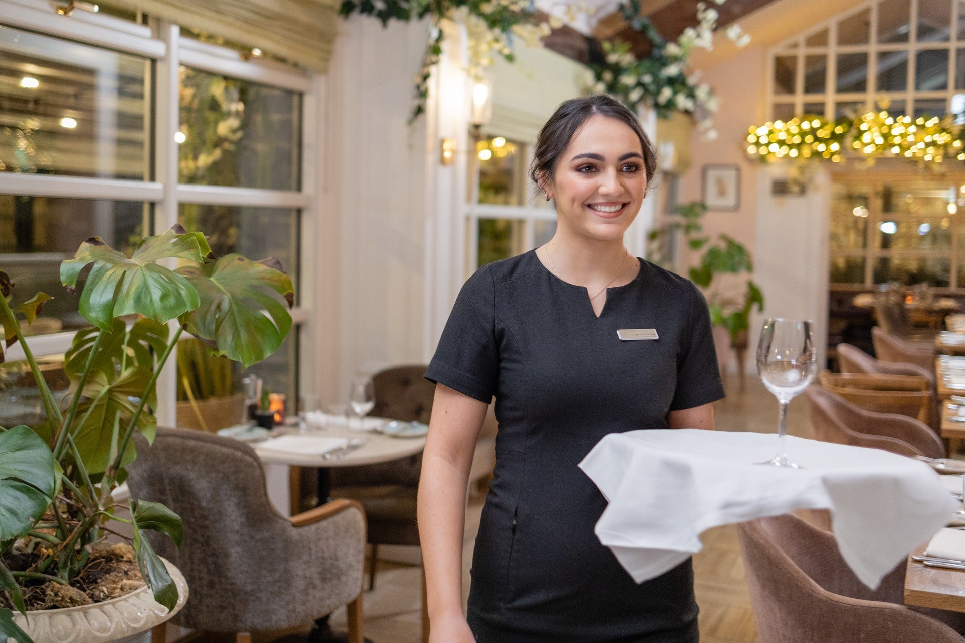 Hospitality staff member in uniform smiling while smoothing a white tablecloth on a set table in a bright restaurant