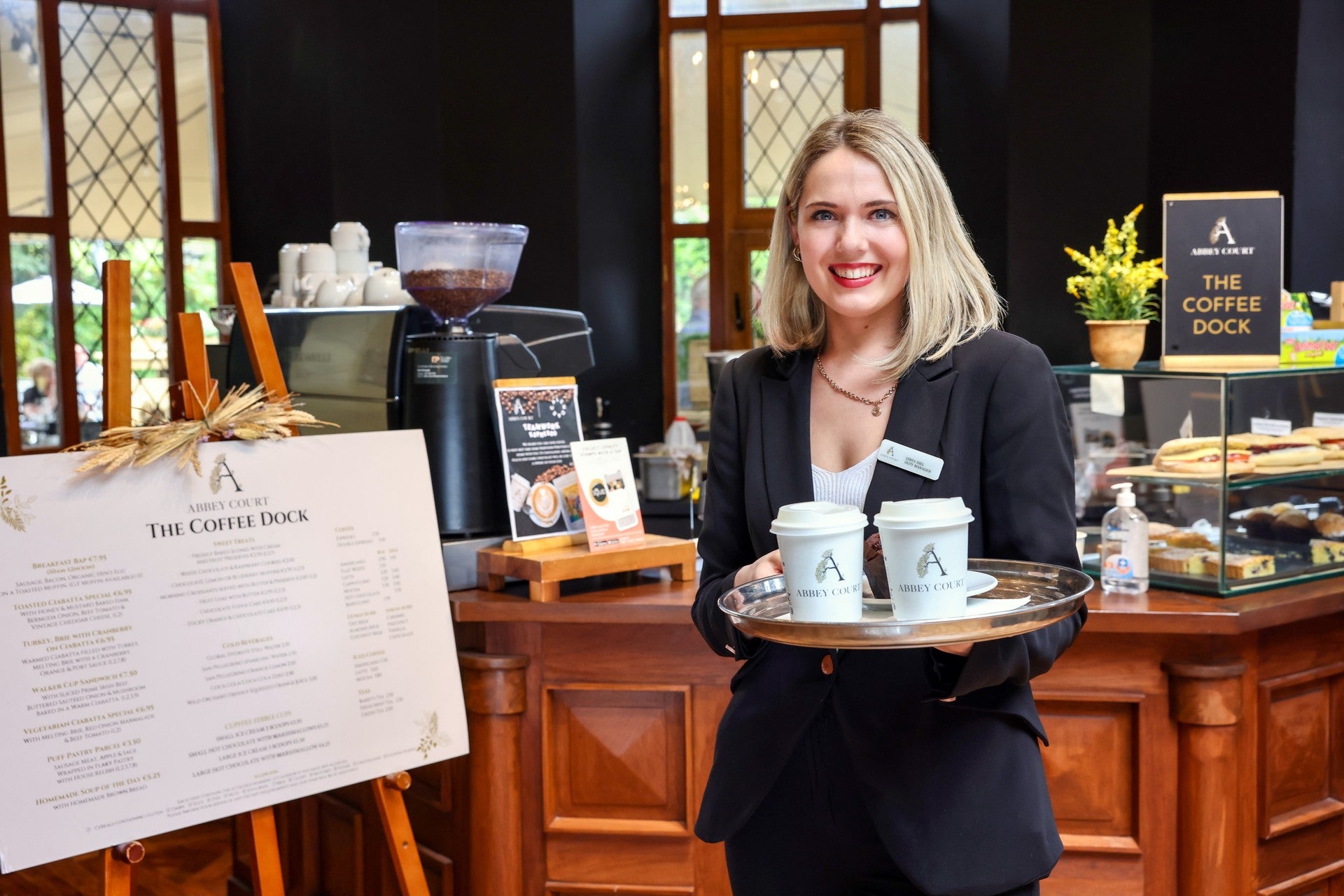 Smiling hotel staff member holding a tray of branded takeaway coffees at a café counter with menu board