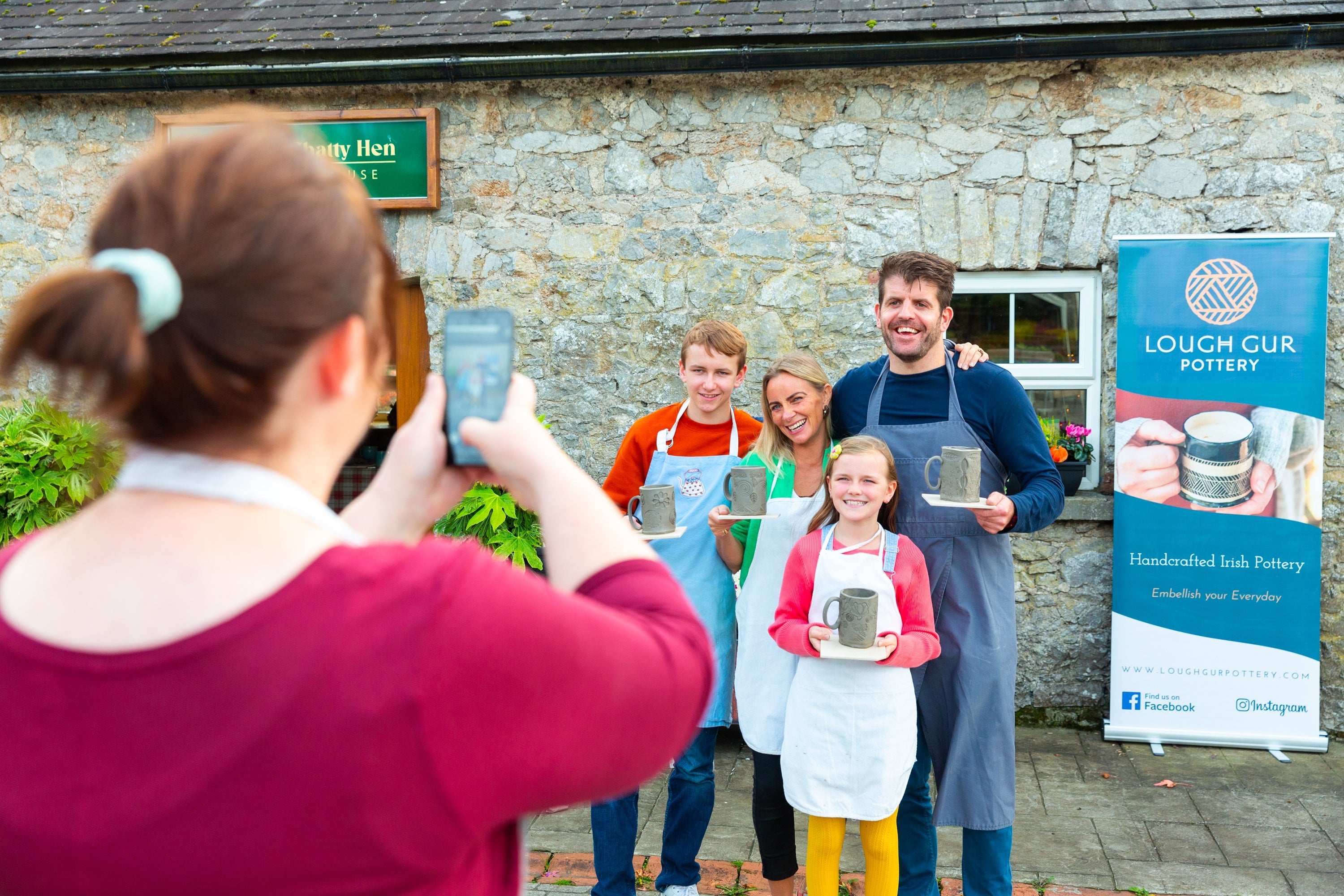 Person using a smartphone to photograph two attendees posing in front of a Fáilte Ireland branded event backdrop