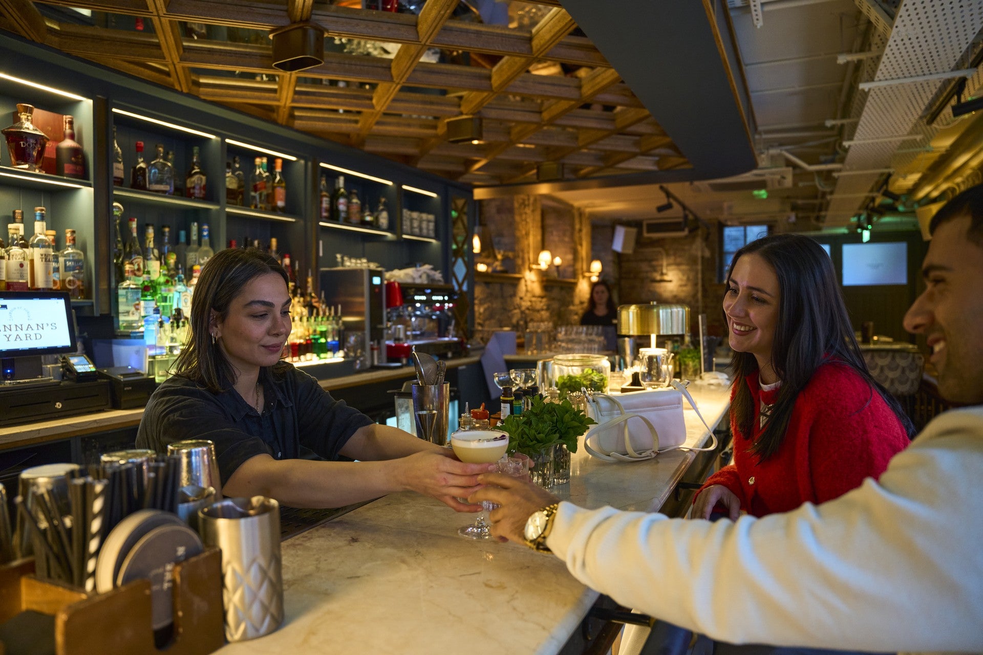 Bar staff member passing a cocktail across a marble bar counter to smiling customers