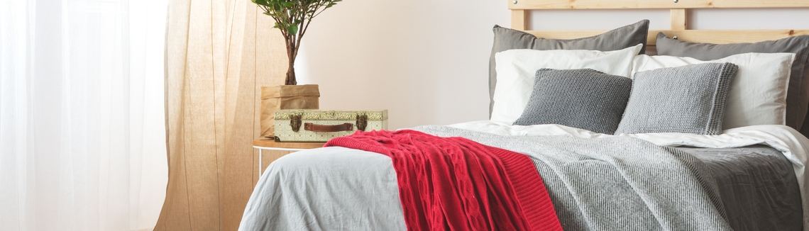 podwójne łóżko z szarą pościelą i leży na nim czerowny pleciony koc