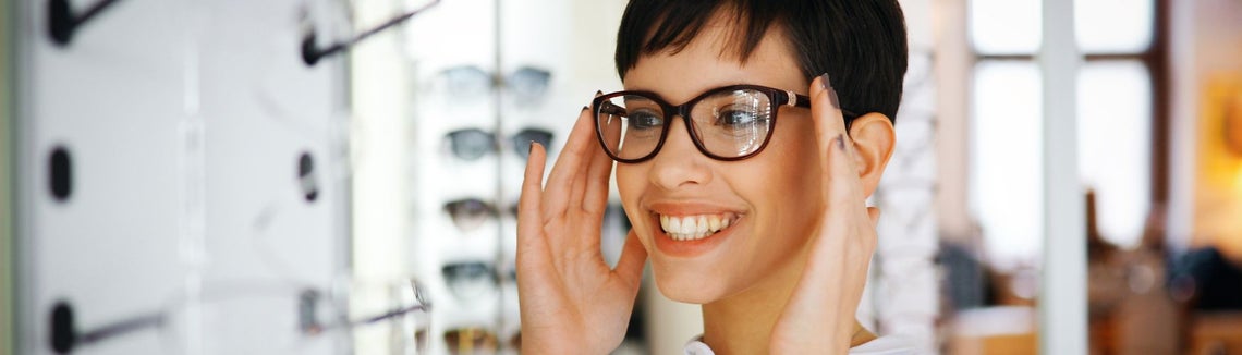 Młoda kobieta przymierza w salonie optycznym okulary z ciemnymi oprawkami. Uśmiecha się.