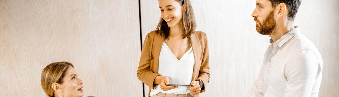 Troje młodych ludzi rozmawia ze sobą w jasnym dobrze oświetlonym pomieszczeniu. Jedna z osób siedzi. 