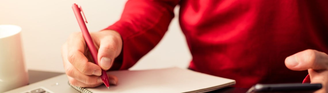 osoba w czerwonym swetrze notuje czerwonym długopisem, w drugiej ręce telefon.