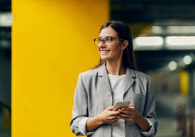 Młoda uśmiechnięta kobieta w okularach, szarym garniturze ze smartfonem