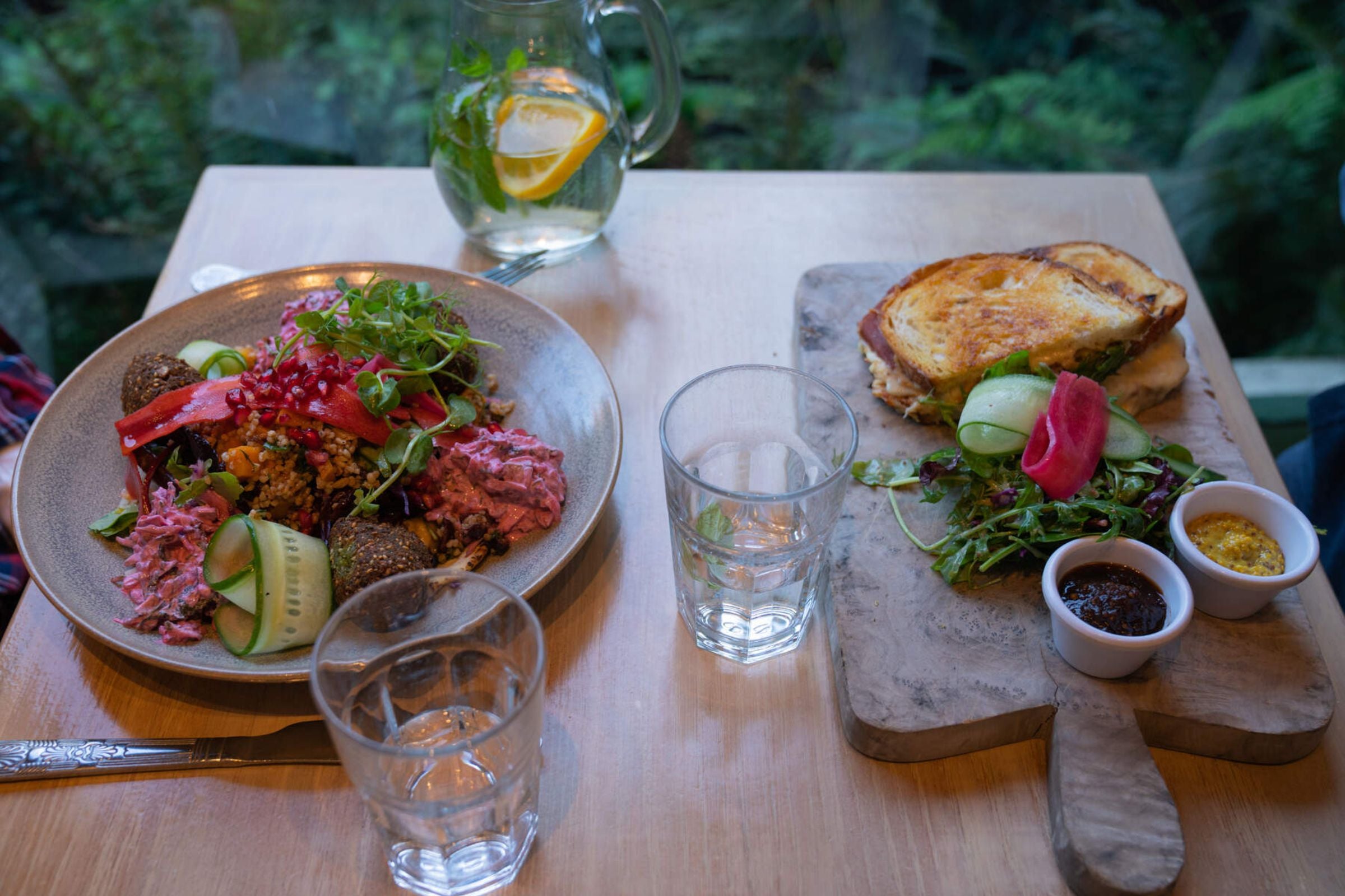 Two plates full of food; one a salad with cucumber and beets, the other a toasted sandwich, all served with lemon water.