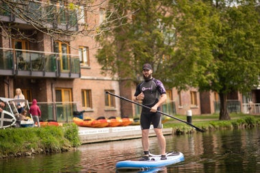 Man in a wetsuit standing on a blue paddle board holding an oar on a canal