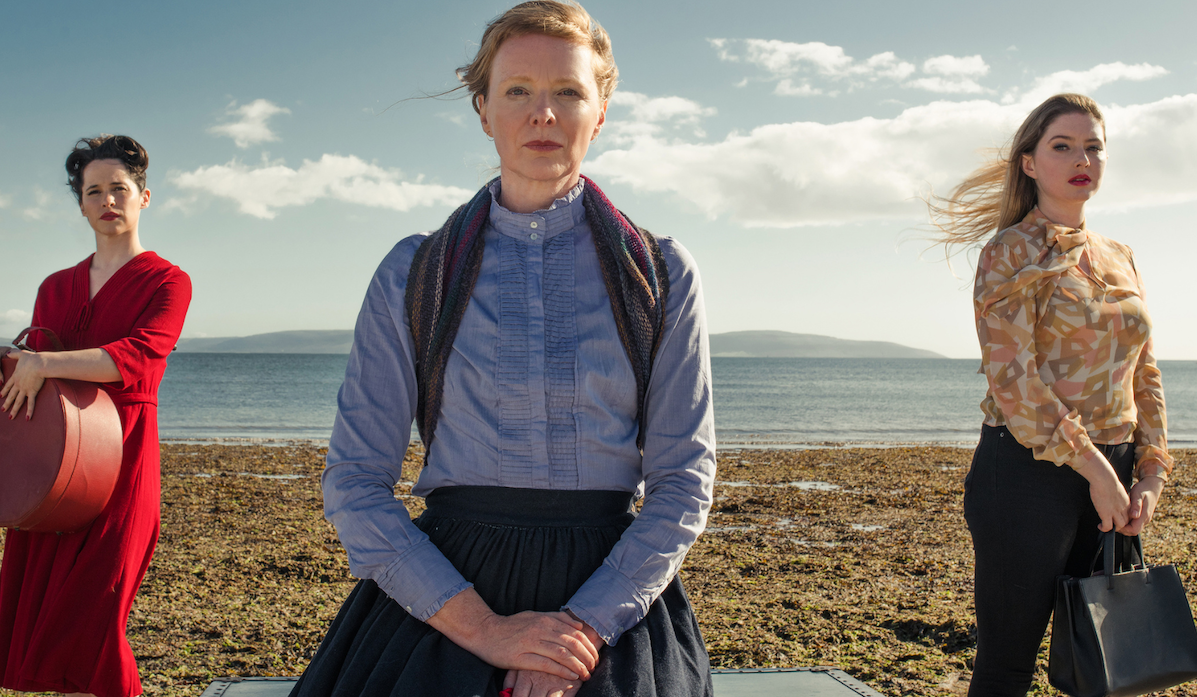 3 women standing apart on a beach with sea in background.