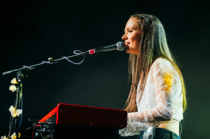 A woman is singing seated at a keyboard with a mic on a long arm in front of her.