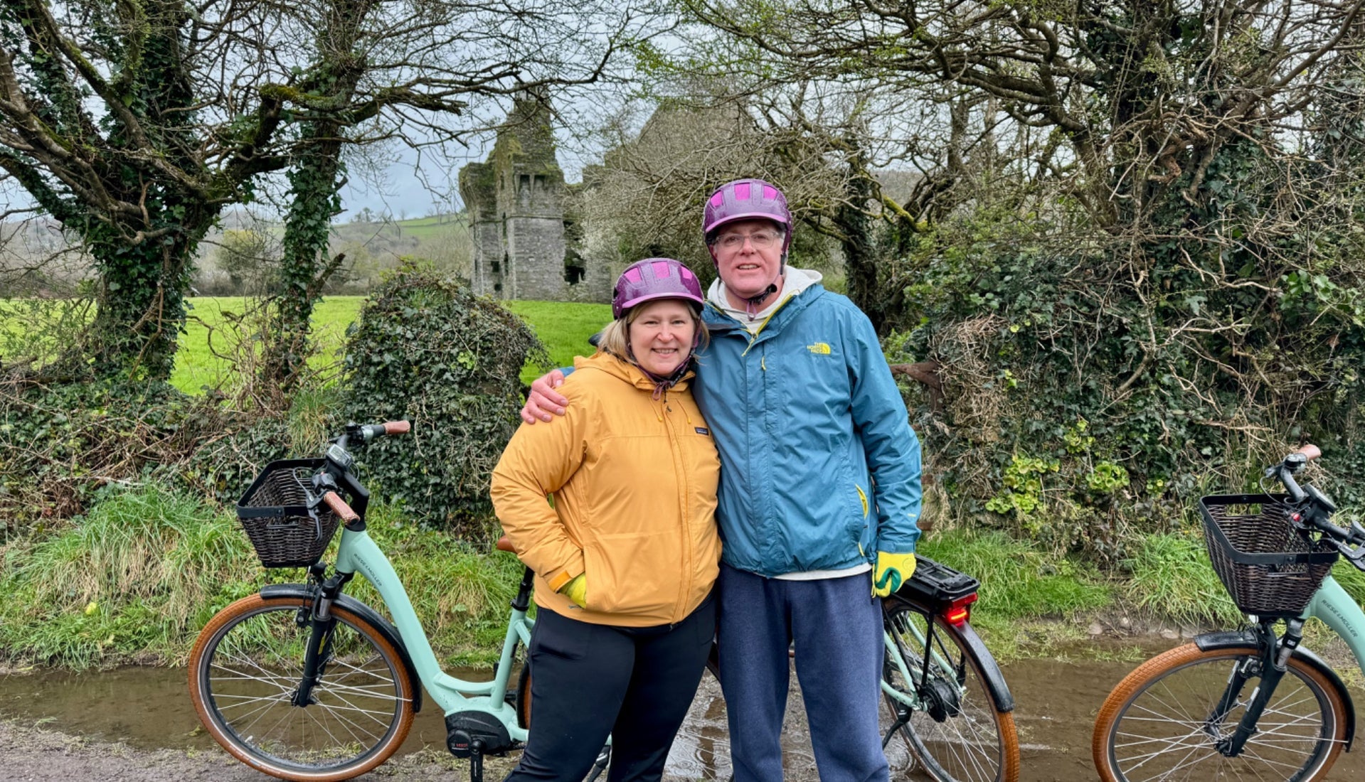 A couple pose for a photo with their eBikes on a country lane near an old ruin