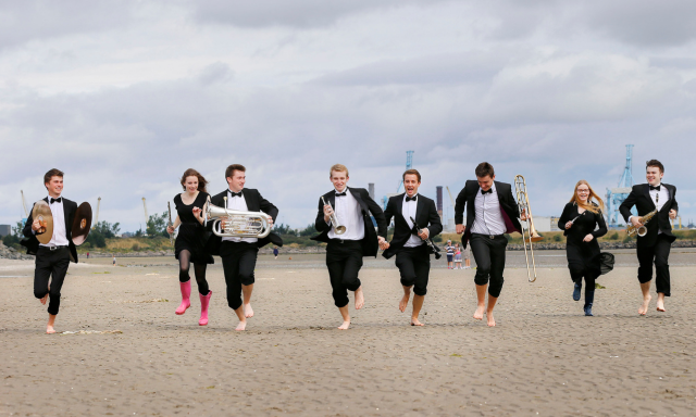 musicians running on Dollymount Strand with the Poolbeg Towers and blue skies behind them