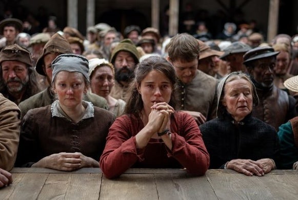 A worried woman resting her elbows on wooden stage at the front of crowd of sad looking people in period costumes.