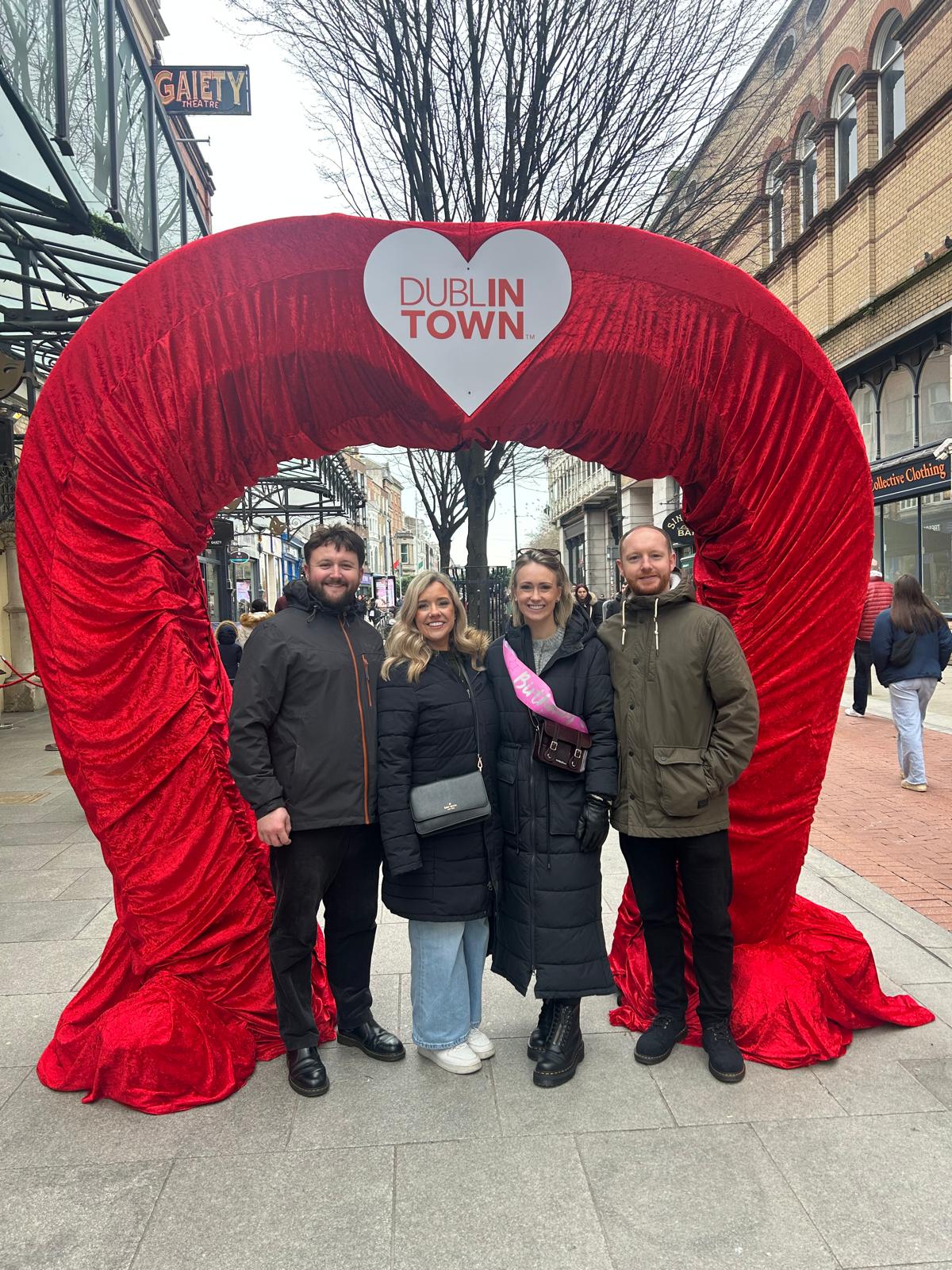 Friends pose beneath a giant red velvet heart arch in the heart of Dublin, part of the city's playful embrace of romance and love around Valentine's season, with the Gaiety Theatre visible in the background.