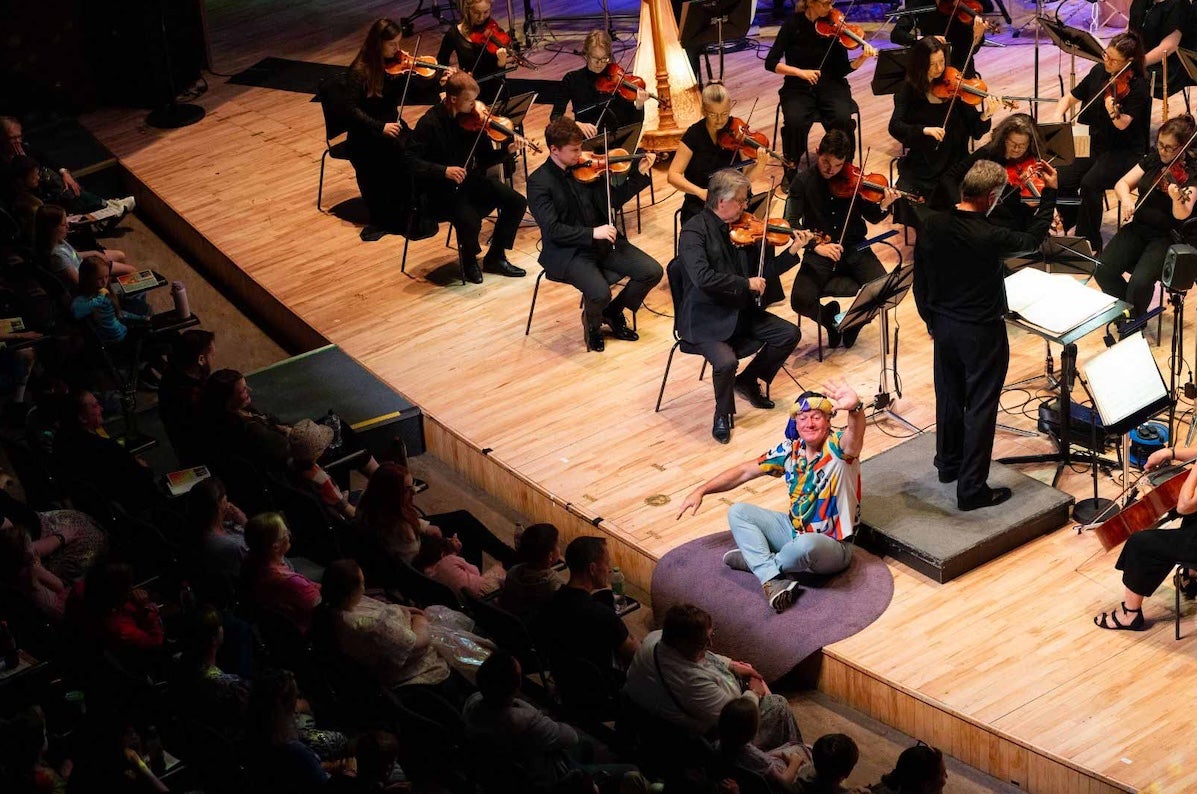 A smiling man in a colourful shirt is seated on the edge of a stage waving at the audience with an orchestra playing behind him.