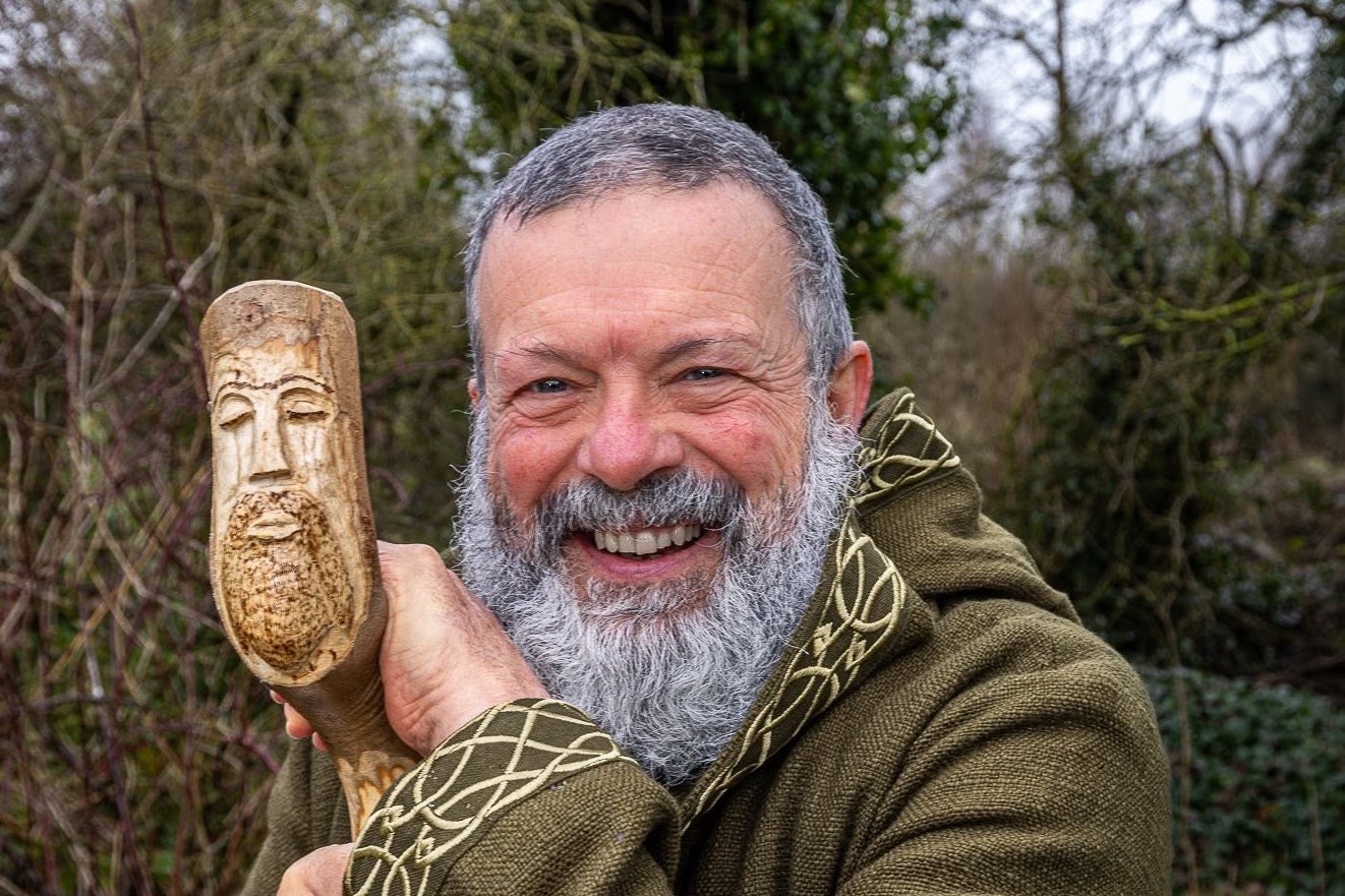 A smiling man leaning on a tall carved stick with the head of a man at the top, outside in winter with bare trees.