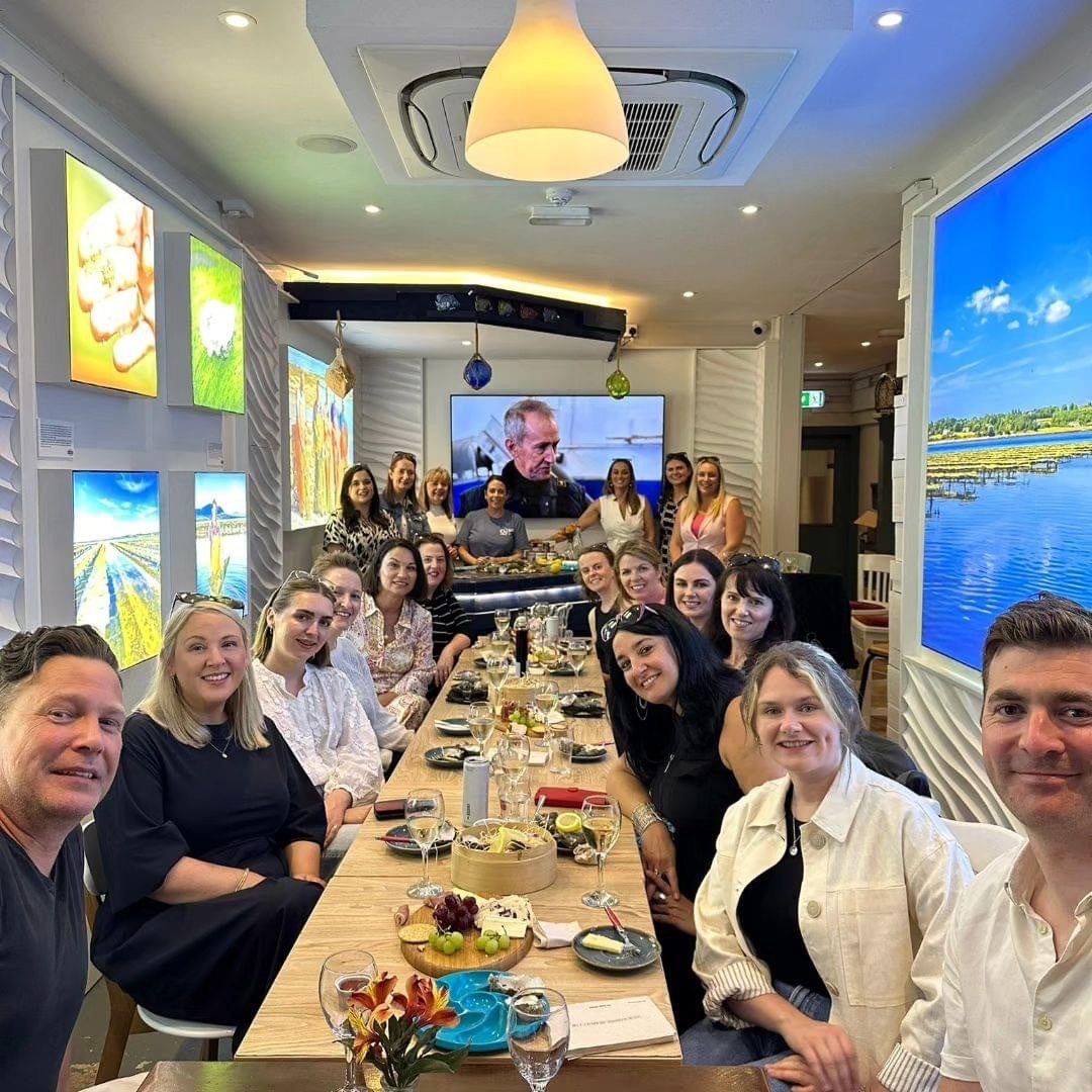 Group of people sitting at a long wooden table with food and wine glasses and bright screens on the walls