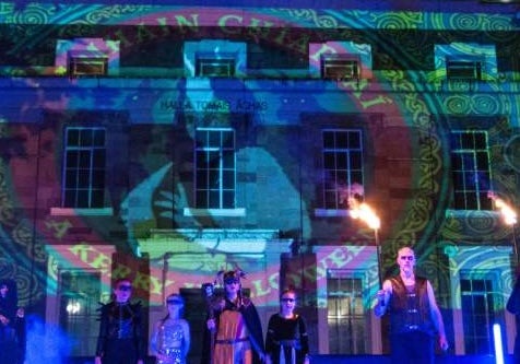 Samhain Chiarraí Street Spectacle, a line of people at night in costumes standing in front of a building lit up with logo from light show.