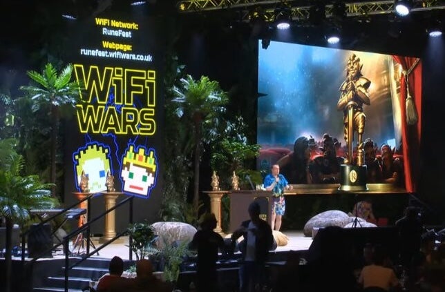 WiFi Wars: Interactive Comedy Game Show, view of a stage with a man talking, a large screen to one side.