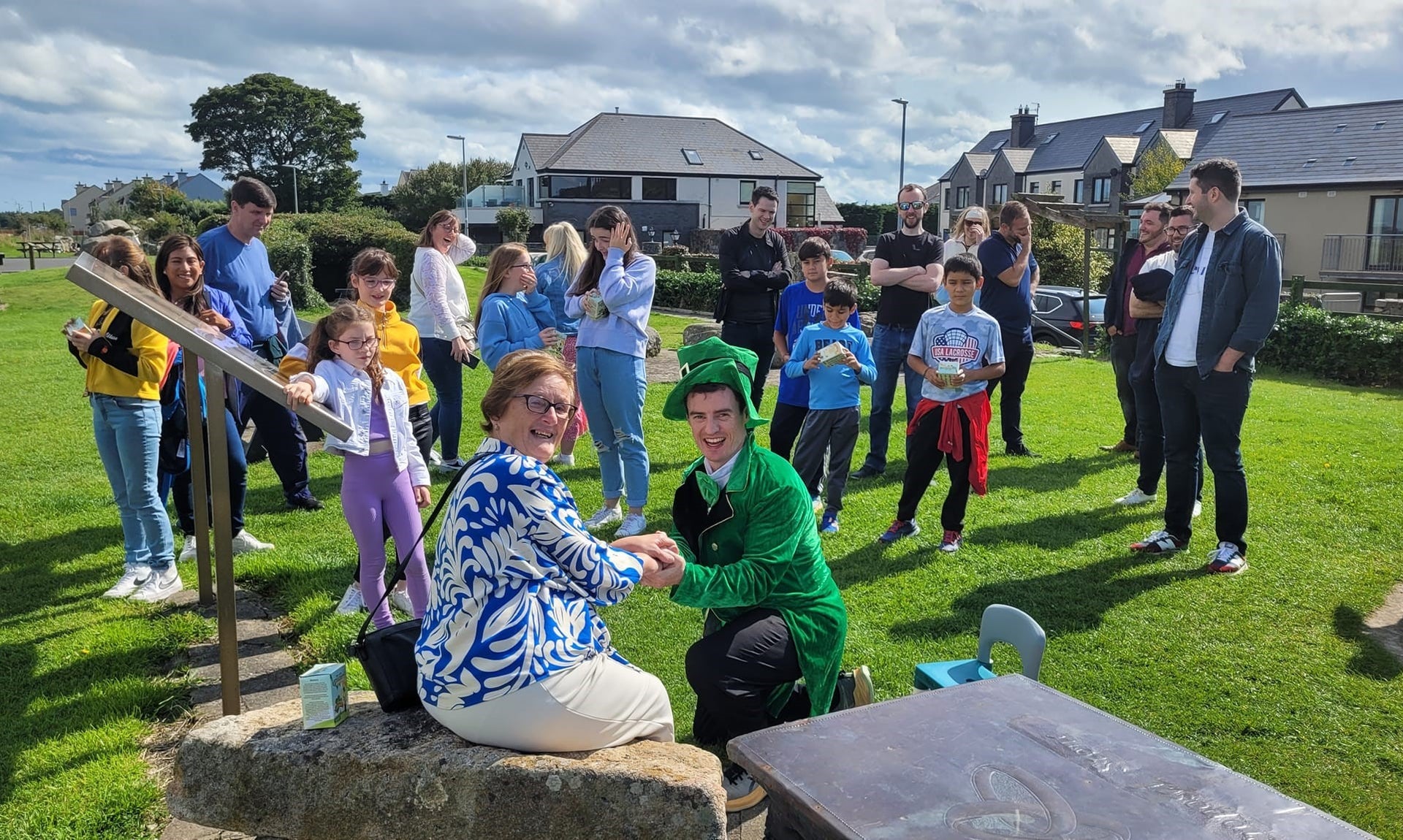 Lady sitting on a rock with a man in a leprechaun costume kneeling down and people in the background