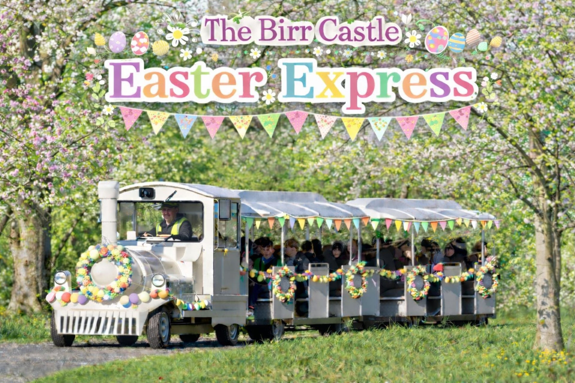 The Birr Castle Easter Express - a white train on a road pulling 2 decorated carriages in eggs and flowers in a woodland setting with blossom.