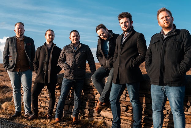 A group of 6 men in coats are standing in a line on the edge of a road with bogland behind.