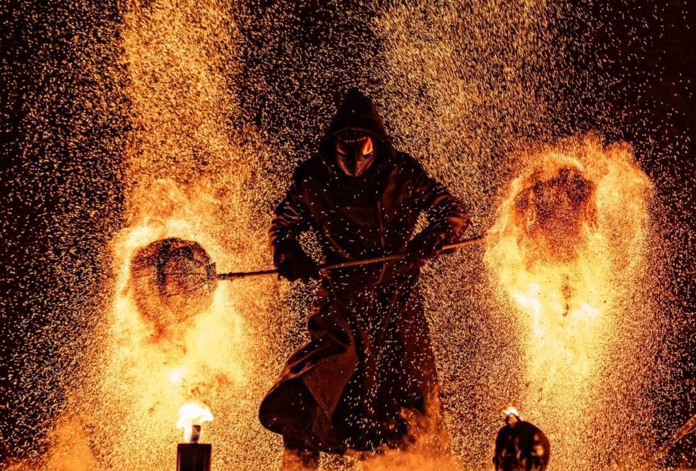 A dark hooded figure against background of flames and sparks from burning.