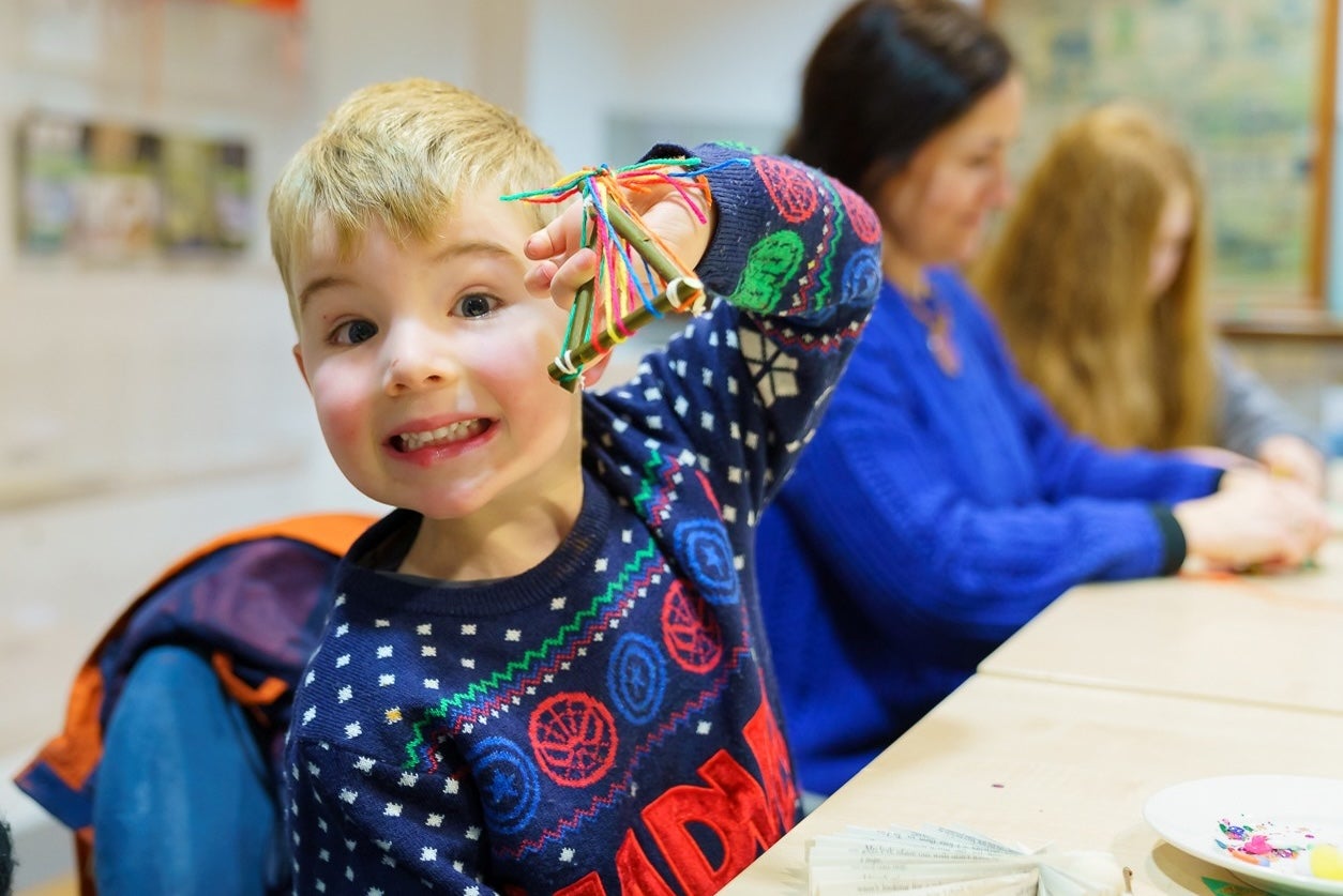 A smiling young boy in a Christmas jumper seated at a table holding up a small handmade decoration