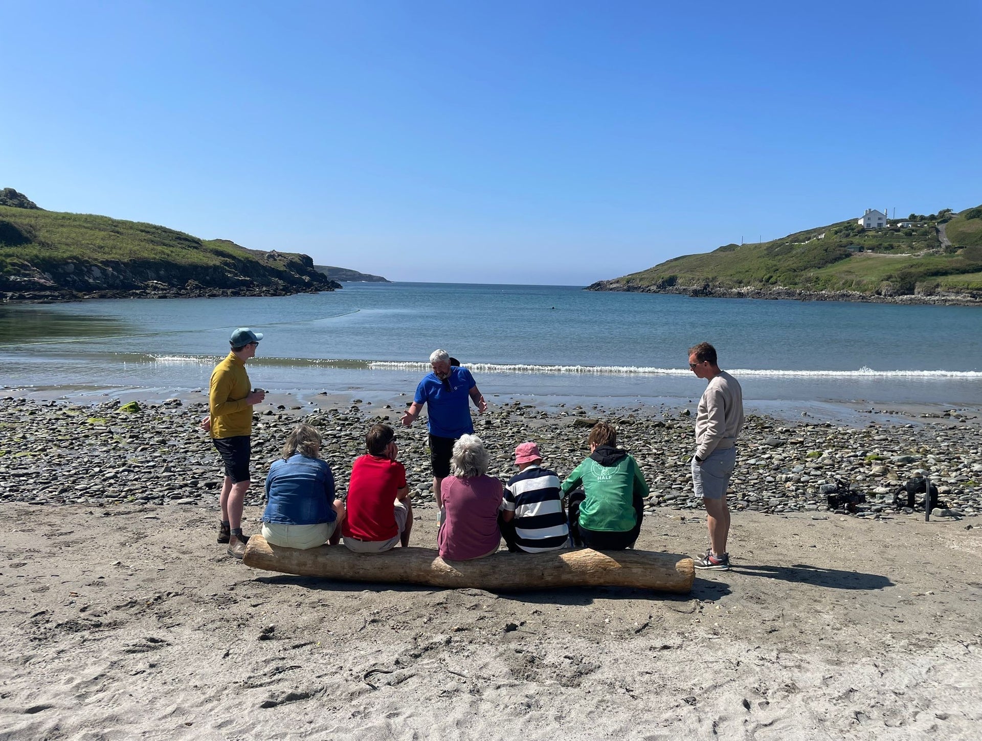 A group of people on a hiking tour sit on a log on a beach listening to their tour guide