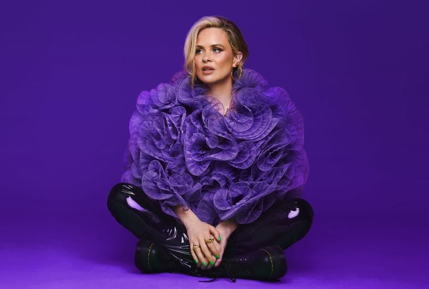 A woman is seated crossed legged on a purple floor wearing a huge, frilly purple top and dark shiny trousers.