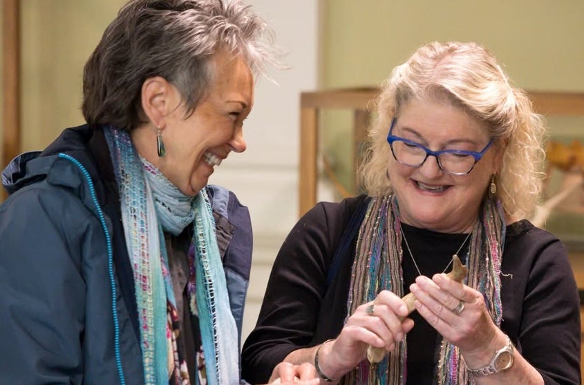 2 laughing women, one holding an artefact with display cases behind.