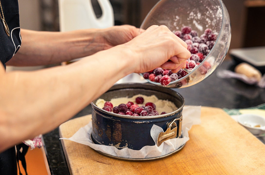 View of pair of hands holding a bowl of red berries, tipping them into a round, deep cake tin