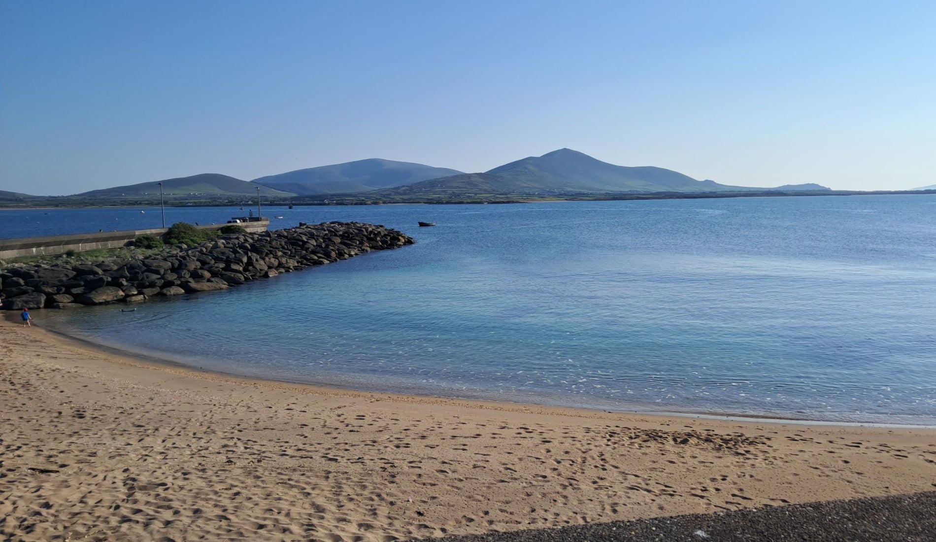A sandy beach beside clear blue water with distant mountains under a bright sky