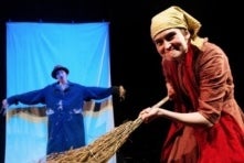 A woman acting as an old woman sweeping with a large twig broom.