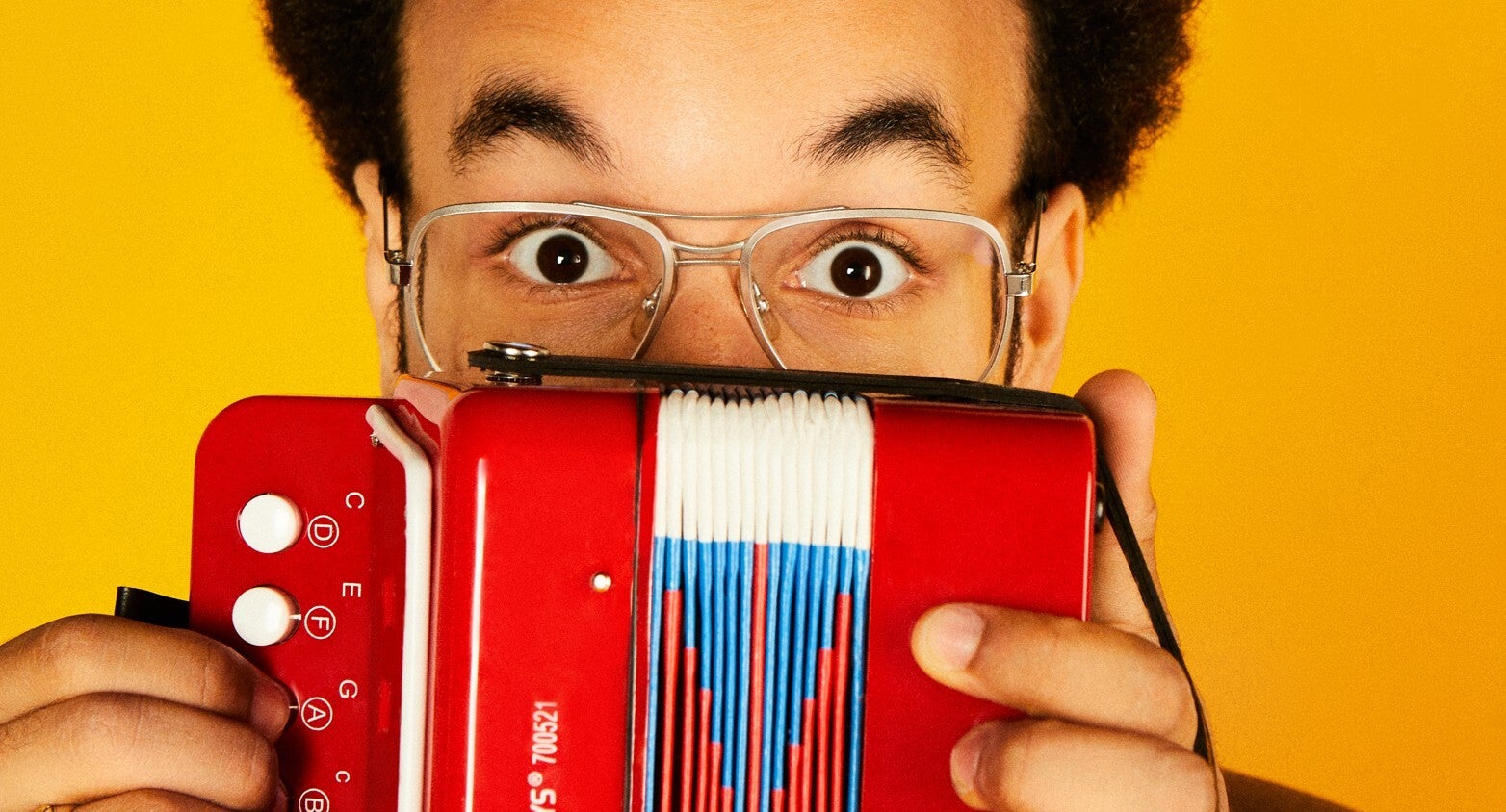 A man in glasses with raised eyebrows peering over the top of a small accordion he's holding up in front of him. 