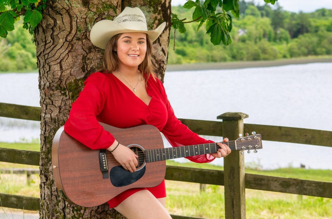 Effie Neill Dance - Gleneagle Arena, a smiling woman playing a guitar, leaning against a tree.