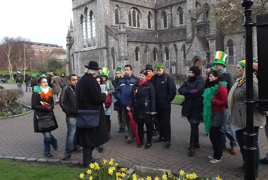 A man in dark overcoat and hat talking to a group of people outside a large, grey stone church.