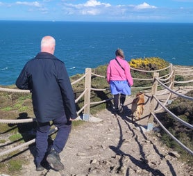 Two people walking a dog along a fenced coastal path overlooking the ocean on a sunny day