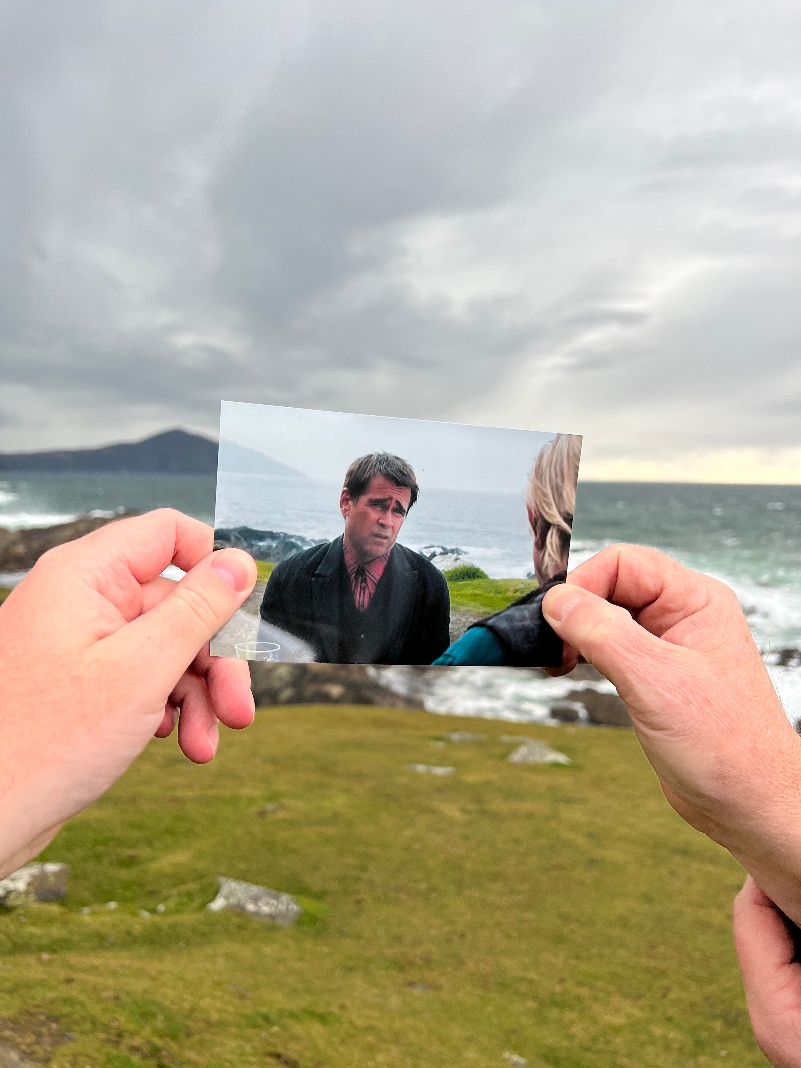 Two hands holding up a picture of a scene from the film, Banshees of Inisherin, that was filmed in that location. The scene pictures actor Colin Farrell expressing a sad face.