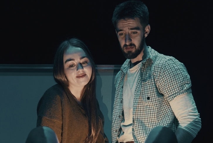 A woman and slightly anxious looking man looking down at something obscured from the viewer, dimly lit.
