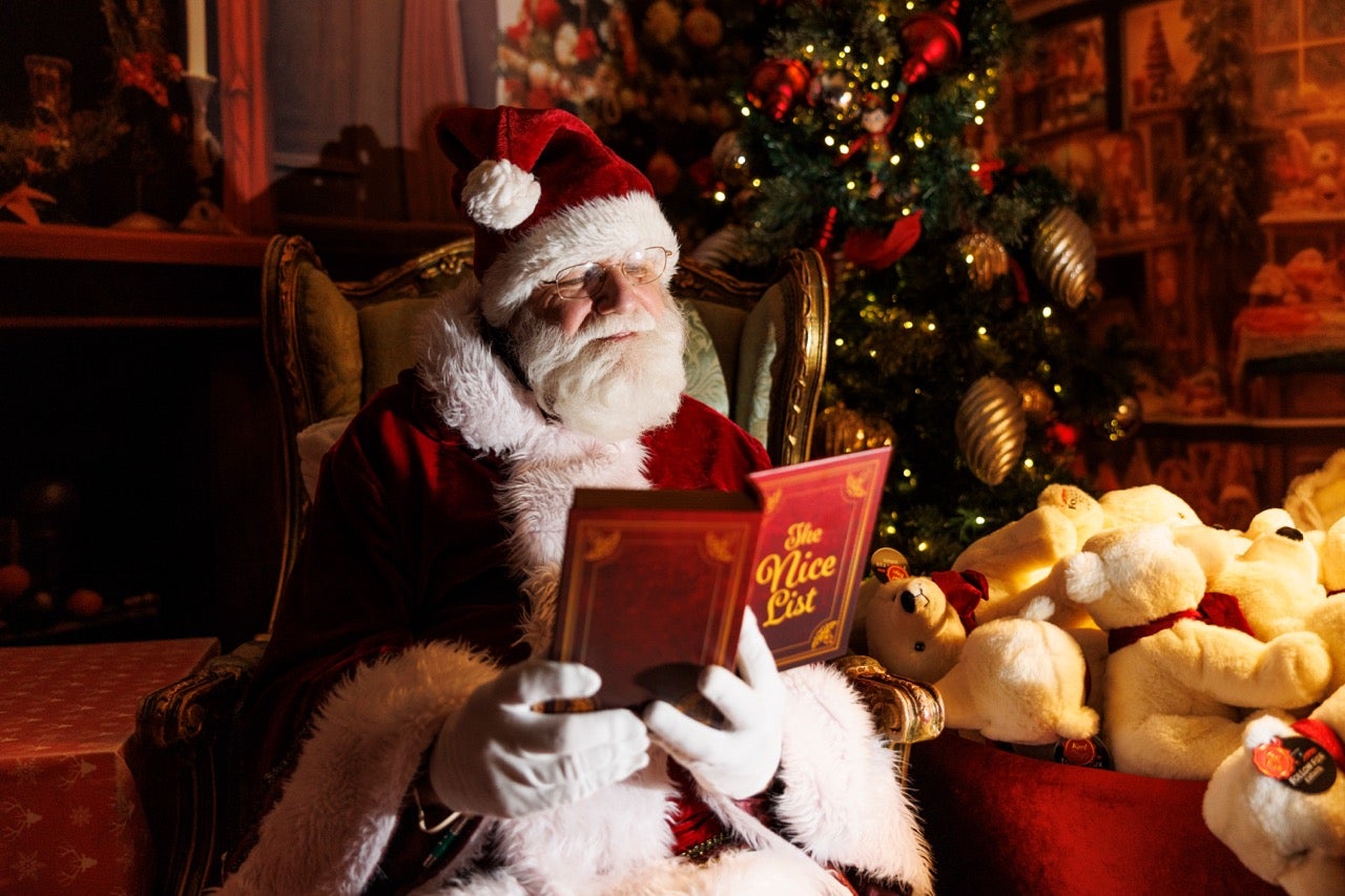 A santa dressed in dark red and white is reading a book with pile of stuffed toys beside him.