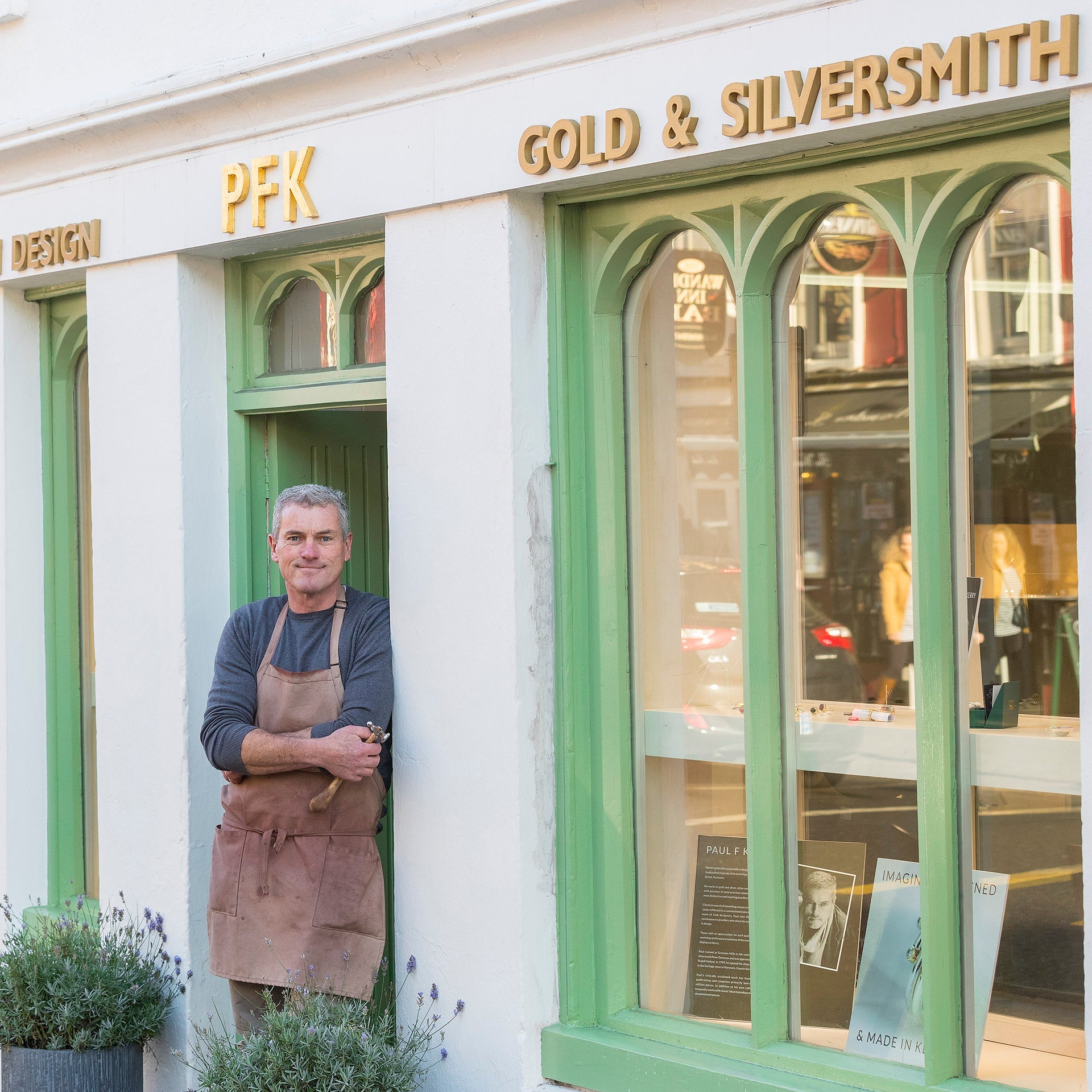 Man in a navy tee shirt and apron leaning against a shop doorway with windows to the right and pfk gold and silversmith written above