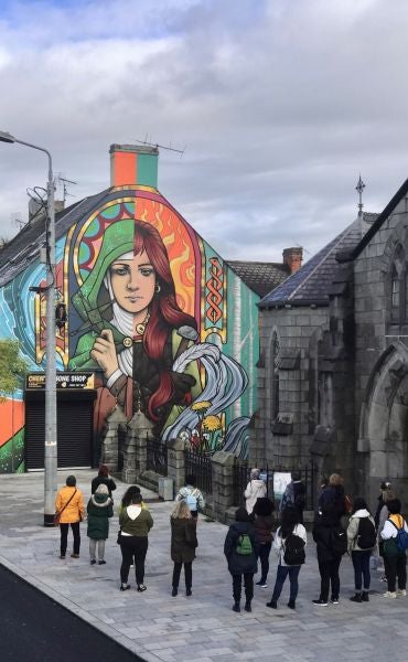 Cruinniú na nÓg 2026 Walking With Legends Live, a group of people on a pavement looking up at the end of building covered in a mural of a woman.