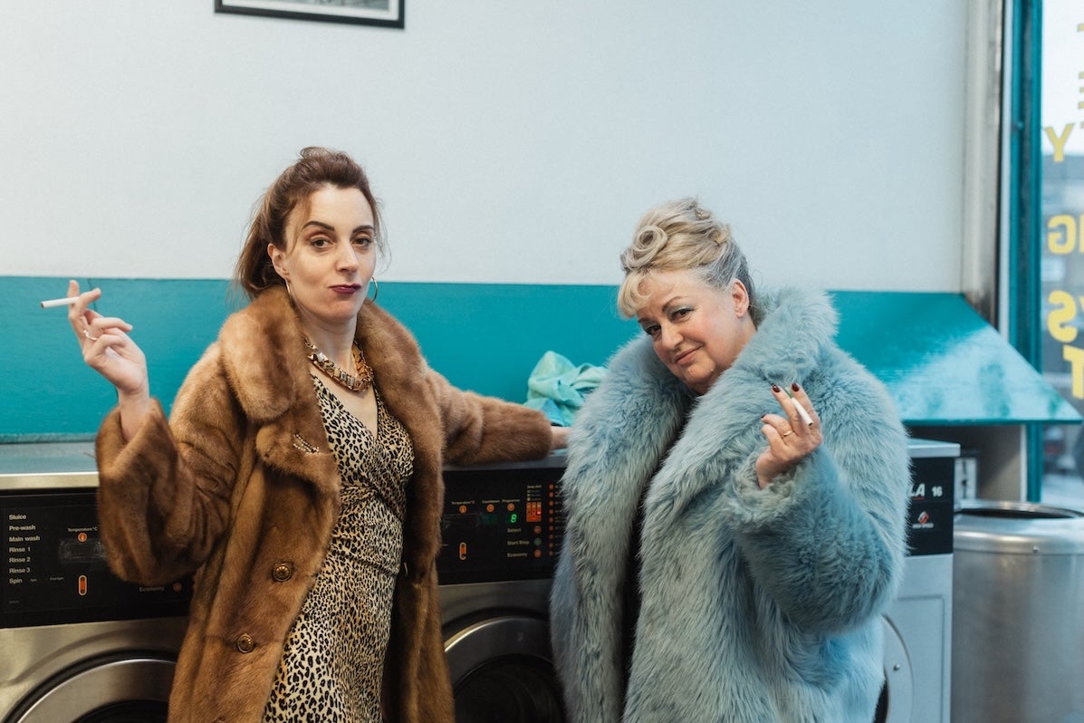 2 women in fur coats are holding cigarettes, leaning against washing machines in a launderette.