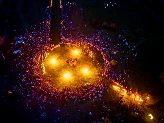 Viewed from above at night, a large crowd gathered around a large circle containing 4 fires.