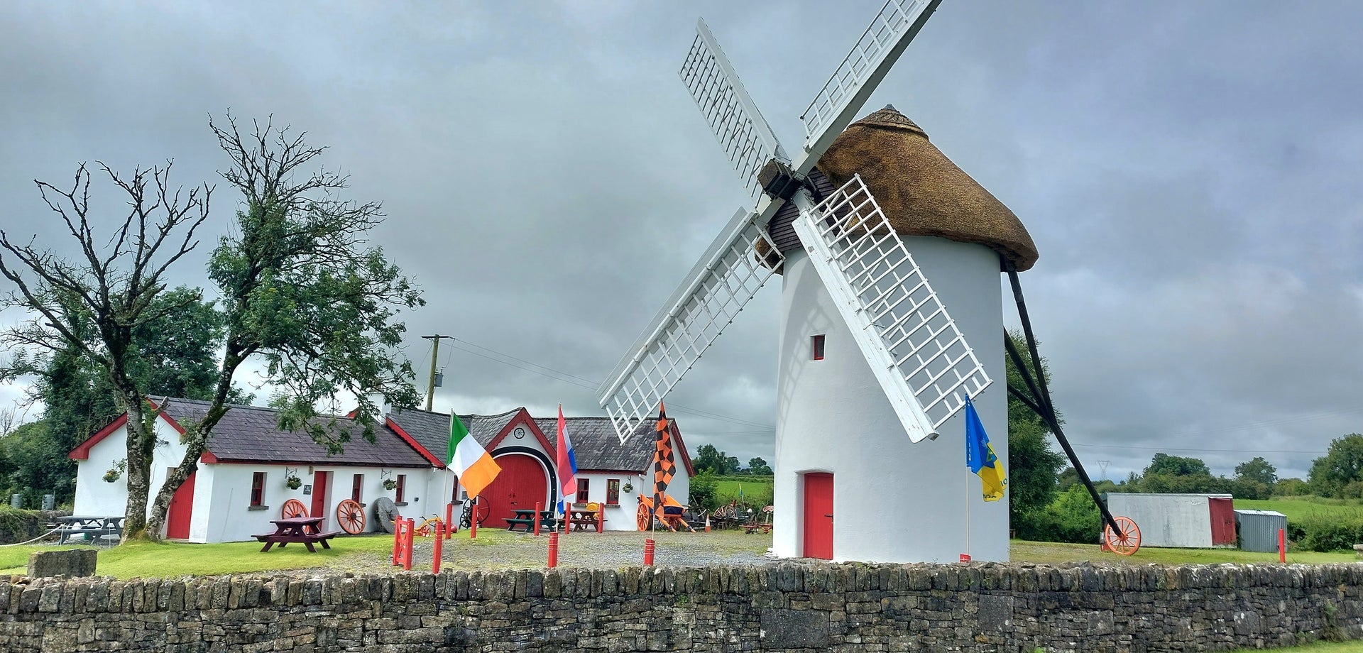 The exterior of the restored Elphin Windmill with a thatched roof and red door and the agricultural museum in the background