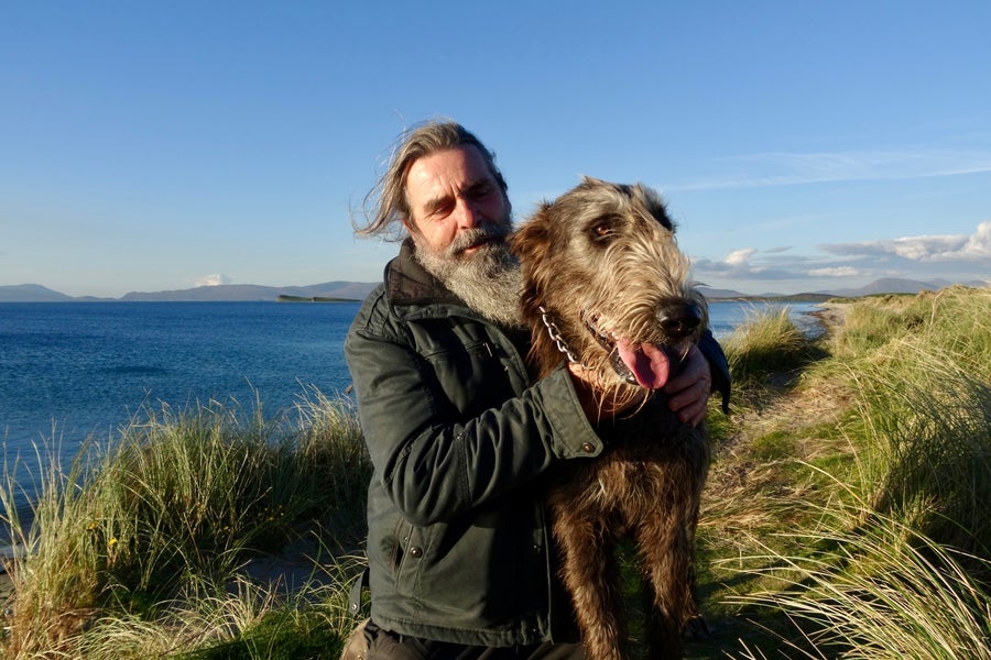 A man posing with an Irish wolfhound in tall grass beside the sea