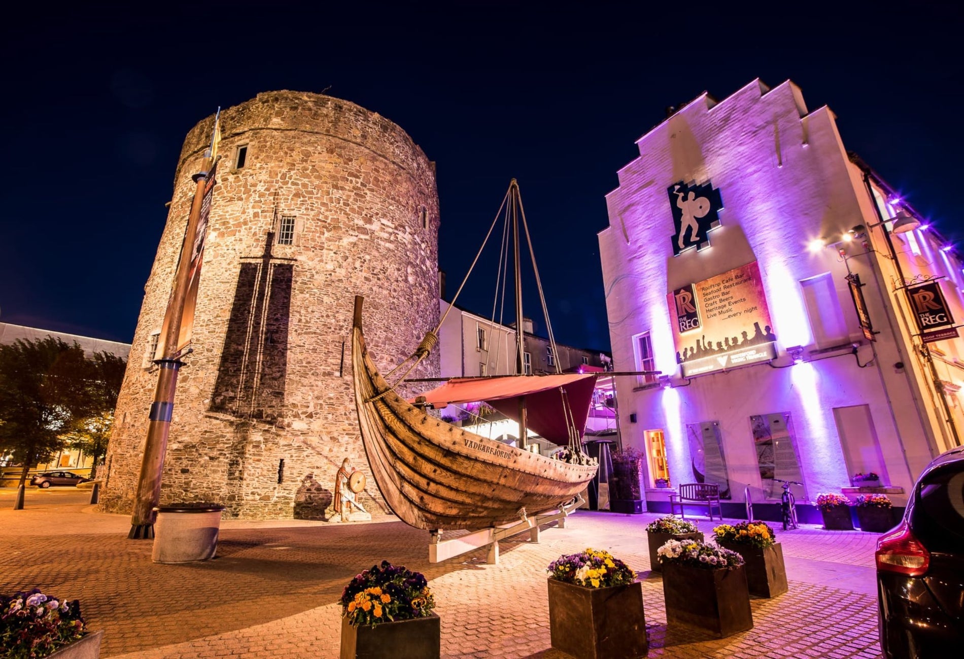 Outside The Reg Waterford lit up at night with pink lights next to Reginald's Tower and a wooden ship