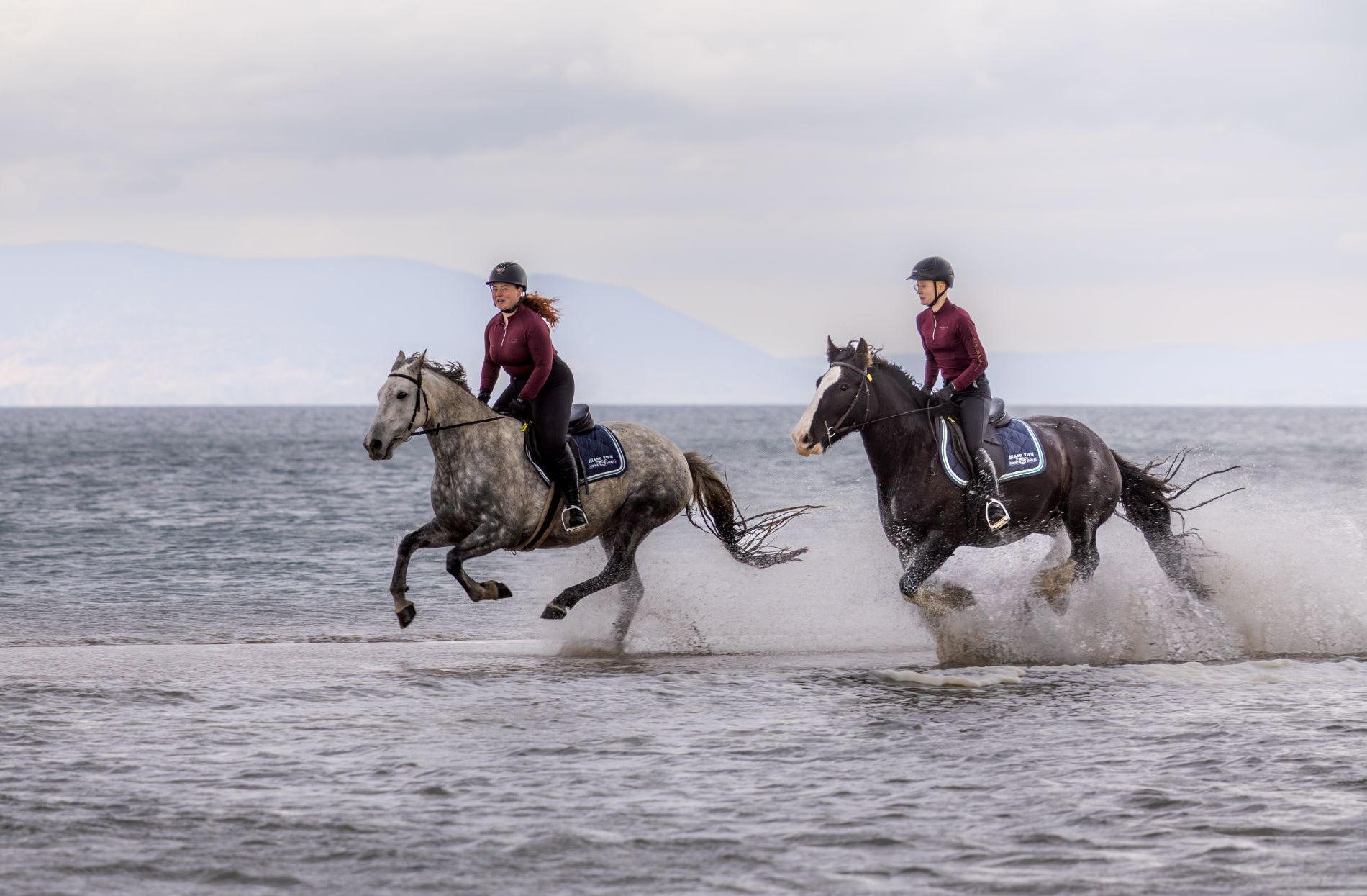 Two people wearing helmets horse riding through water